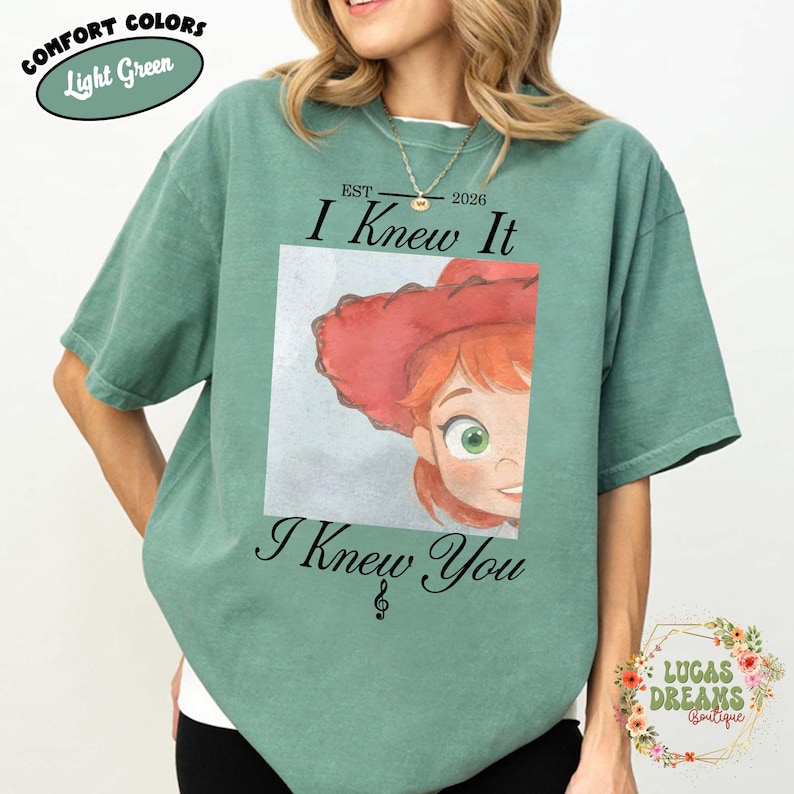 May include: A light green short-sleeve t-shirt featuring a graphic print of a cartoon character with red hair and a red hat. The text above the graphic reads EST 2026 I Knew It and the text below reads I Knew You with a small musical note symbol.