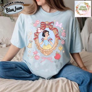 以下が含まれることがあります： 金色のフレームに白雪姫のグラフィックが描かれた水色のTシャツ。ピンクのバラ、リボン、赤いリンゴで囲まれています。「Comfort Colors」と「Blue Jean」の文字がプリントされています。画像には「LUCAS DREAMS BOUTIQUE」の文字も含まれています。