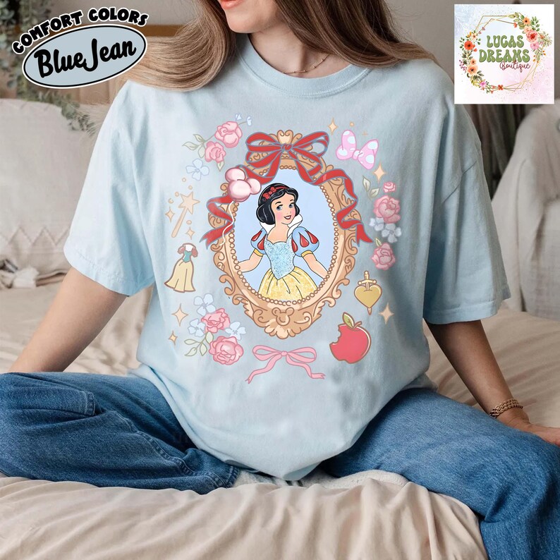 以下が含まれることがあります： 金色のフレームに白雪姫のグラフィックが描かれた水色のTシャツ。ピンクのバラ、リボン、赤いリンゴで囲まれています。「Comfort Colors」と「Blue Jean」の文字がプリントされています。画像には「LUCAS DREAMS BOUTIQUE」の文字も含まれています。
