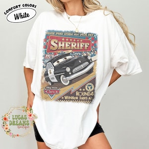 May include: White t-shirt featuring a graphic of Sheriff from Disney Pixar's Cars. The design includes the text "SHERIFF", "RADIATOR SPRINGS SATURDAY NIGHT SPECIAL RACE", and "ROUND 4 WINNER TAKES ALL". The shirt has a relaxed fit.