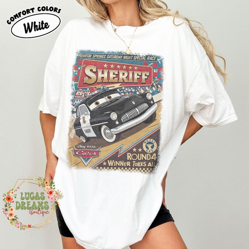 May include: White t-shirt featuring a graphic of Sheriff from Disney Pixar's Cars. The design includes the text "SHERIFF", "RADIATOR SPRINGS SATURDAY NIGHT SPECIAL RACE", and "ROUND 4 WINNER TAKES ALL". The shirt has a relaxed fit.