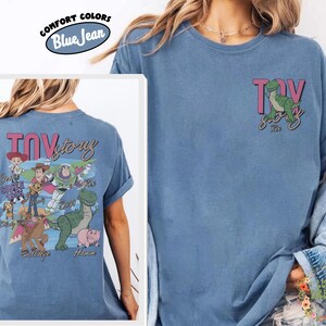 Peut inclure: T-shirt bleu jean avec un motif du film "Toy Story". Le devant pr&eacute;sente Rex le dinosaure, et le dos un motif plus grand avec des personnages et le mot "Toy Story" en rose. Marque Comfort Colors.