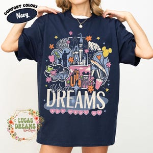 K&ouml;nnte beinhalten: Marineblaues T-Shirt mit einem farbenfrohen Grafikdesign mit ikonischen Wahrzeichen und Figuren. Der Text "What Dreams Are Made Of" ist deutlich sichtbar. Das Shirt tr&auml;gt auch die Aufschrift "Comfort Colors" und "Navy".