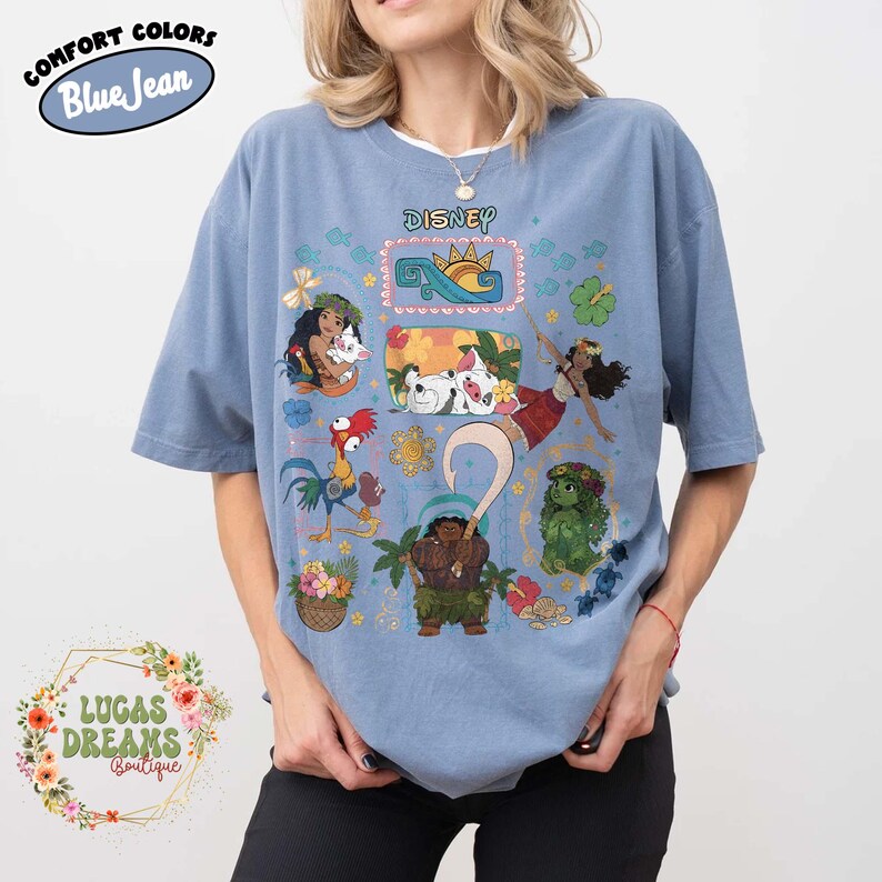 May include: Pale blue short-sleeved t-shirt with a Disney graphic featuring characters from the movie Moana. The shirt has a crew neck and a relaxed fit. The text "Comfort Colors Blue Jean" is on the top left.
