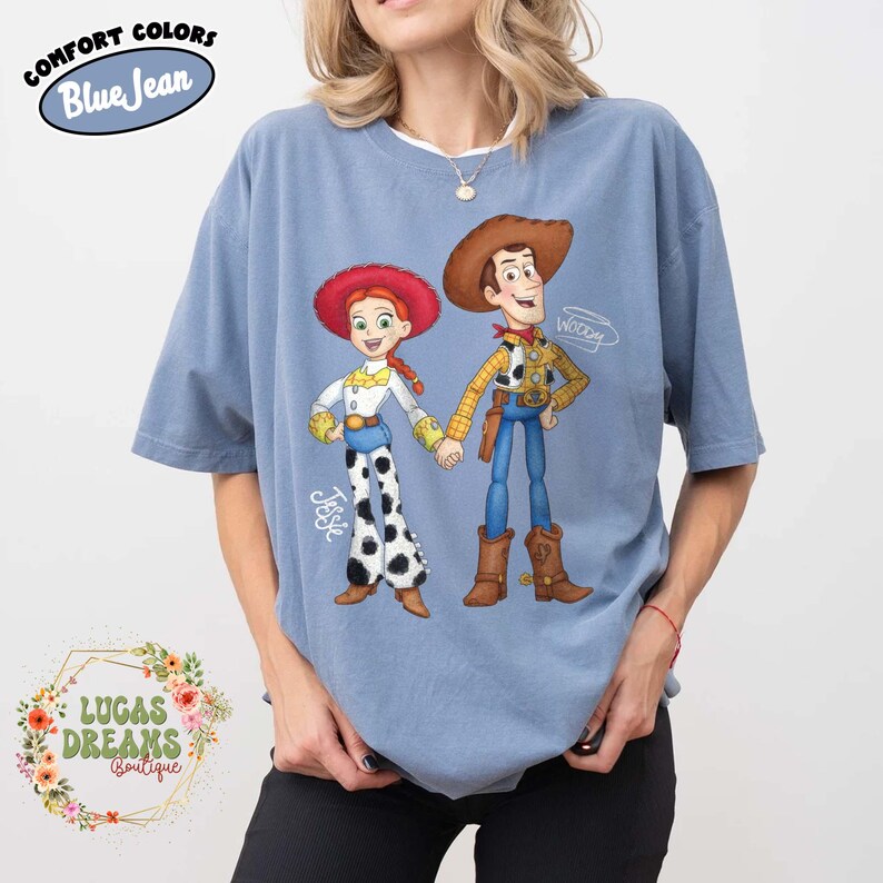 Puede incluir: Camiseta azul claro con un dise&ntilde;o de los personajes de Toy Story, Jessie y Woody, tomados de la mano. La camiseta tiene el texto "Comfort Colors Blue Jean" y "Lucas Dreams Boutique".