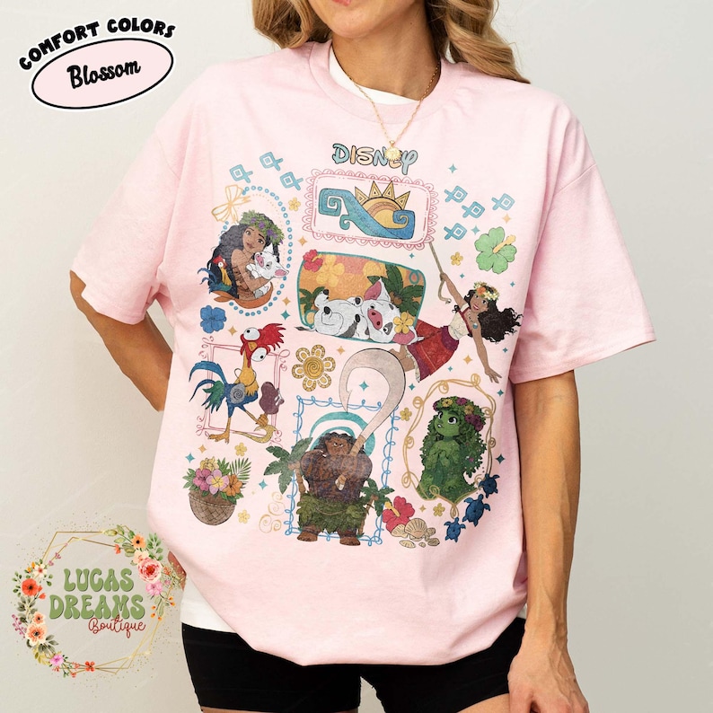 May include: Pale pink t-shirt featuring a Disney graphic with characters from the film Moana. The design includes Moana, Maui, Pua, Hei Hei, and other characters. The shirt has a crew neck and short sleeves, with the word "Disney" printed on it.