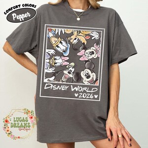 Op de afbeelding: Een grijs T-shirt met een Disney World-afbeelding met Mickey Mouse, Minnie Mouse, Donald Duck en Goofy. De afbeelding is omlijst in een witte rechthoek, met "Disney World 2026" eronder gedrukt. Het shirt heeft bovenaan de tekst "Comfort Colors Pepper".