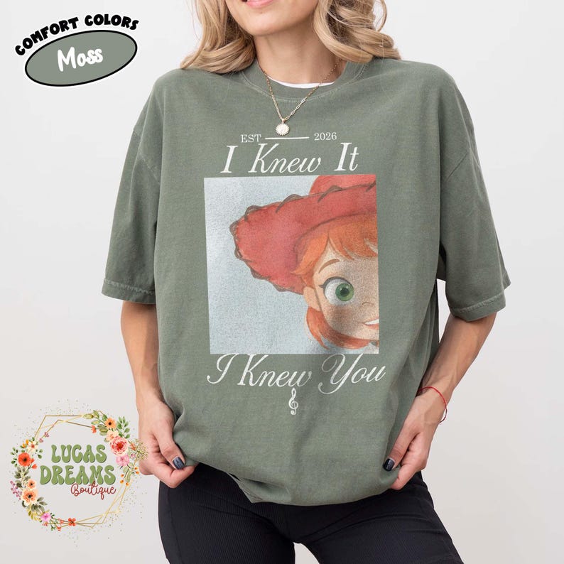 May include: A moss green short-sleeve t-shirt featuring a graphic print of a red-haired character wearing a red hat. The text reads EST 2026 I Knew It I Knew You with a small musical note symbol. The shirt has a crew neckline and a relaxed fit.