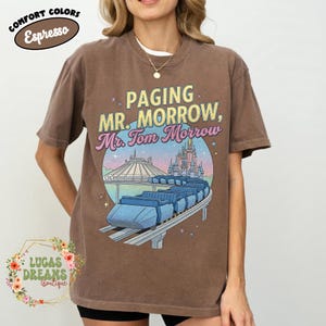 May include: Brown t-shirt featuring the text "Paging Mr. Morrow, Mr. Tom Morrow" and a graphic of a futuristic ride with a castle and space mountain in the background. The shirt also has the words "Comfort Colors Espresso" in a circle.