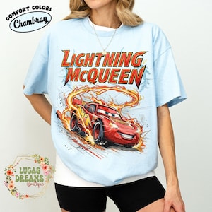 May include: Light blue t-shirt featuring Lightning McQueen from the Cars movie. The graphic shows the red race car with flames and the text "LIGHTNING McQUEEN". The shirt has the text "COMFORT COLORS Chambray" and "LUCAS DREAMS Boutique".