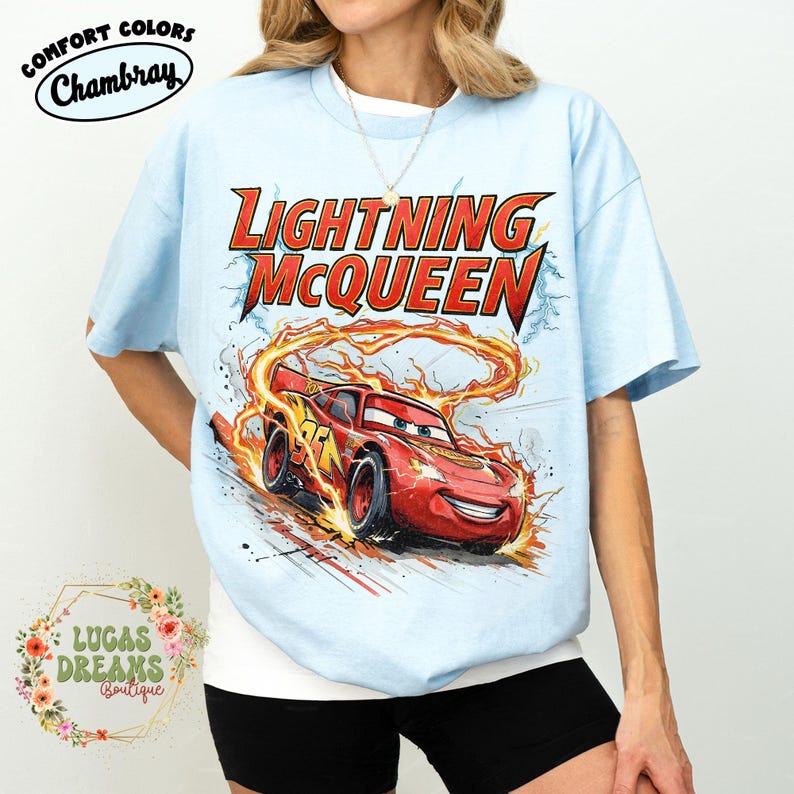 May include: Light blue t-shirt featuring Lightning McQueen from the Cars movie. The graphic shows the red race car with flames and the text "LIGHTNING McQUEEN". The shirt has the text "COMFORT COLORS Chambray" and "LUCAS DREAMS Boutique".