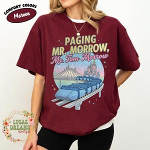 May include: Maroon t-shirt featuring the text "Paging Mr. Morrow, Mr. Tom Morrow" and a graphic of a roller coaster and a castle. The shirt also has the words "Comfort Colors" and "Lucas Dreams Boutique".