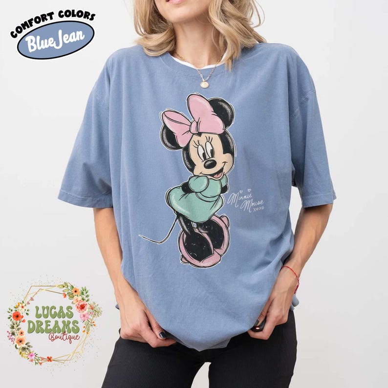 May include: A blue jean colored t-shirt featuring a graphic of Minnie Mouse wearing a pink bow and a light green dress. The text "Minnie Mouse xoxo" is printed in white next to the character. The shirt has a crew neckline and short sleeves.