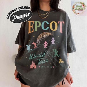 Puede incluir: Camiseta gris oscuro con la palabra "EPCOT" en letras coloridas. La camiseta presenta un gr&aacute;fico de la esfera EPCOT, personajes de Disney y las palabras "World Tour" y "Est. 1982". La camiseta est&aacute; etiquetada como "Comfort Colors Pepper".