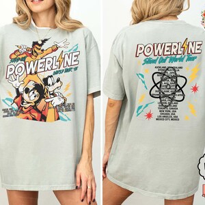 Puede incluir: Camiseta verde claro con un gr&aacute;fico de personajes de dibujos animados y la palabra "POWERLINE". La parte trasera de la camiseta enumera ciudades de una gira mundial. La camiseta tiene un corte holgado.