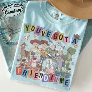以下が含まれることがあります： トイ・ストーリーの様々なアニメキャラクターのグラフィックプリントが施された、ライトブルーのクルーネックTシャツ。キャラクターグループの上下には、「YOU'VE GOT A FRIEND IN ME」というテキストがカラフルなブロック体で描かれています。