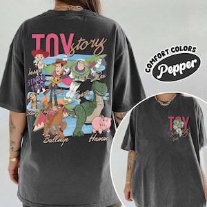 Peut inclure: T-shirt gris fonc&eacute; avec un motif Toy Story repr&eacute;sentant Woody, Buzz l'&Eacute;clair et Rex. Le design comprend les mots "Toy Story" en rose et "Depuis 1995". Le t-shirt est un Comfort Colors Pepper.