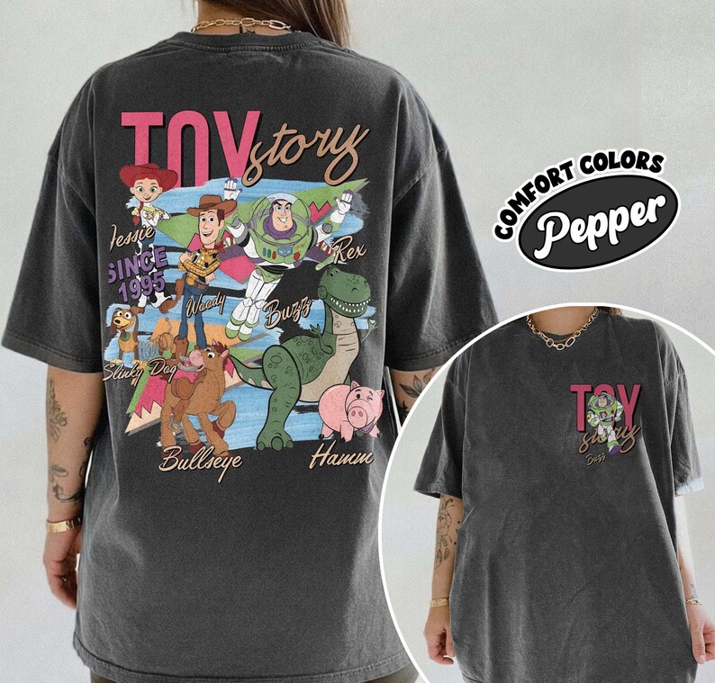Peut inclure: T-shirt gris fonc&eacute; avec un motif Toy Story repr&eacute;sentant Woody, Buzz l'&Eacute;clair et Rex. Le design comprend les mots "Toy Story" en rose et "Depuis 1995". Le t-shirt est un Comfort Colors Pepper.