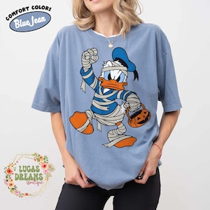 May include: Blue short-sleeved t-shirt featuring a cartoon Donald Duck dressed as a mummy, holding a jack-o'-lantern bucket. The shirt has the words "Comfort Colors Blue Jean" printed on it. The shirt is part of the Lucas Dreams Boutique collection.