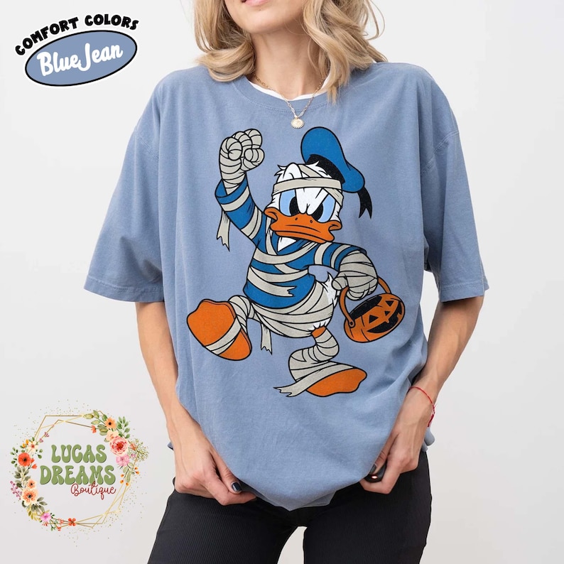 May include: Blue short-sleeved t-shirt featuring a cartoon Donald Duck dressed as a mummy, holding a jack-o'-lantern bucket. The shirt has the words "Comfort Colors Blue Jean" printed on it. The shirt is part of the Lucas Dreams Boutique collection.