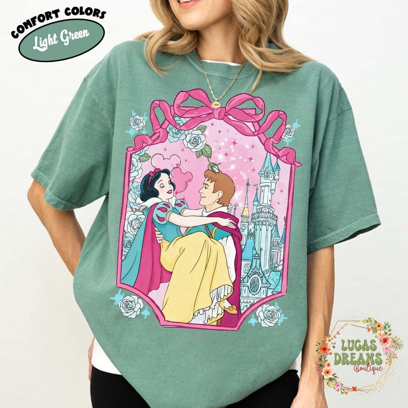 Puede incluir: Camiseta Comfort Colors verde claro con una ilustraci&oacute;n enmarcada de Blancanieves y el Pr&iacute;ncipe Encantador. La ilustraci&oacute;n est&aacute; rodeada de rosas rosas y un lazo rosa. Tambi&eacute;n se ve el texto "Light Green" y "Lucas Dreams Boutique".