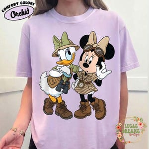May include: A lilac t-shirt featuring Donald Duck and Minnie Mouse in safari outfits. The characters are wearing khaki-coloured safari attire, hats, and boots. The shirt has the words "Comfort Colors" and "Orchid" printed on it.