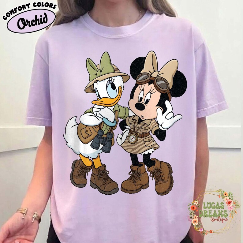 May include: A lilac t-shirt featuring Donald Duck and Minnie Mouse in safari outfits. The characters are wearing khaki-coloured safari attire, hats, and boots. The shirt has the words "Comfort Colors" and "Orchid" printed on it.