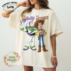 Puede incluir: Camiseta de color marfil con Buzz Lightyear y Woody de Toy Story, con el texto "to infinity and beyond". La camiseta tiene cuello redondo y mangas cortas. El logotipo de la marca Comfort Colors es visible.