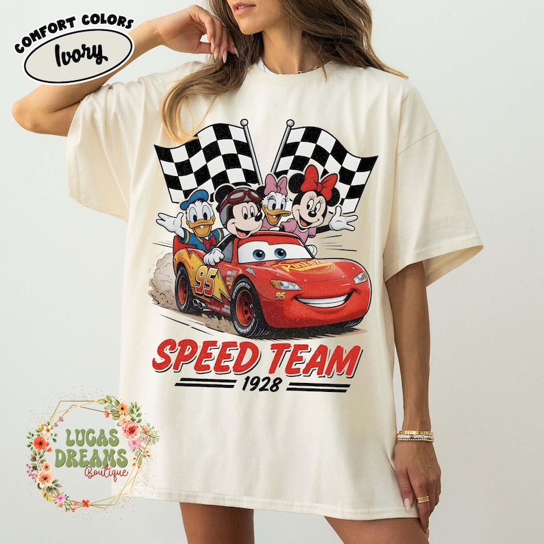 Op de afbeelding: Ivoor kleurig T-shirt met een afbeelding van Disney-figuren in een rode raceauto met geblokte vlaggen. De tekst "SPEED TEAM 1928" staat onder de auto. Het shirt heeft de woorden "COMFORT COLORS" en "Ivory" linksboven.