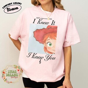 May include: A light pink short-sleeve t-shirt featuring a graphic of a cartoon character with red hair and a red hat. The text "EST 2026 I Knew It I Knew You" is printed in black above and below the graphic, with a musical treble clef symbol.