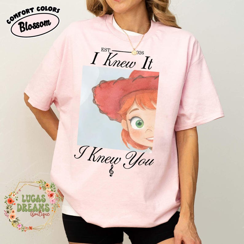 May include: A light pink short-sleeve t-shirt featuring a graphic of a cartoon character with red hair and a red hat. The text "EST 2026 I Knew It I Knew You" is printed in black above and below the graphic, with a musical treble clef symbol.