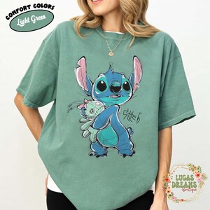May include: A light green short-sleeve t-shirt featuring a graphic illustration of the blue character Stitch holding a small doll. The design includes sketch-like black outlines and the text Stitch written in a stylized script font on the front.