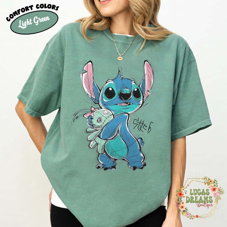 May include: A light green short-sleeve t-shirt featuring a graphic illustration of the blue character Stitch holding a small doll. The design includes sketch-like black outlines and the text Stitch written in a stylized script font on the front.