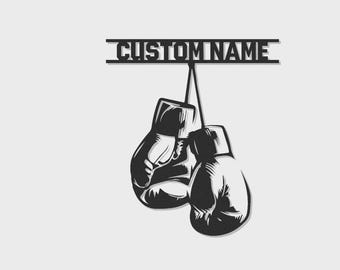 May include: A black metal wall art piece featuring a pair of hanging boxing gloves suspended from a horizontal bar. The bar displays the text CUSTOM NAME in a bold, blocky font. The design is rendered in a solid black silhouette style.