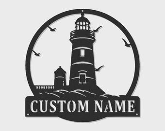 May include: A circular black metal wall sign featuring a lighthouse silhouette, crashing waves, and flying birds. The design includes a rectangular banner at the bottom with the cut-out text CUSTOM NAME, providing a space for personalization.