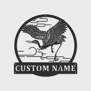 May include: Black metal wall art featuring a crane in flight within a circular frame, with stylized clouds. The bottom of the frame has a rectangular plaque with the text "CUSTOM NAME". The crane is detailed with intricate wing and body patterns.