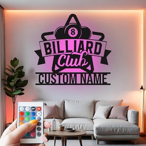 Può includere: Un'opera d'arte da parete in metallo nero con le parole "BILLIARD Club CUSTOM NAME" in un carattere stilizzato. Il design include palle da biliardo e uno stendardo. Un telecomando è tenuto in primo piano, suggerendo l'illuminazione a LED che cambia colore.
