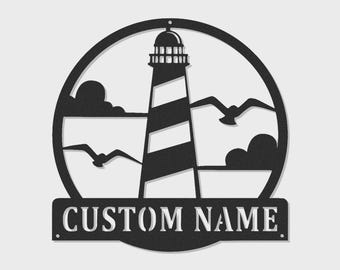 May include: A circular black metal wall sign featuring a lighthouse with diagonal stripes, two flying seagulls, and clouds. Below the circular frame is a rectangular banner with the cutout text CUSTOM NAME, designed for personalization.