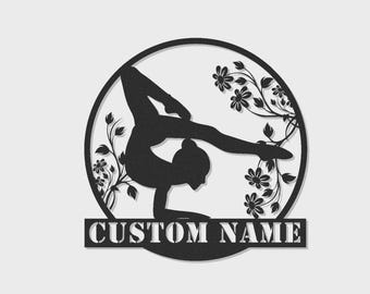 May include: A black circular metal wall art piece featuring a silhouette of a gymnast performing a handstand pose. The design includes floral vine accents on the sides and a rectangular banner at the bottom with the text CUSTOM NAME.