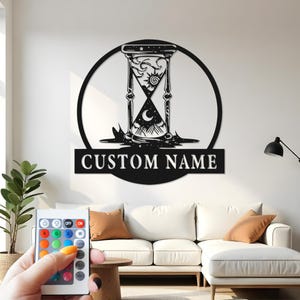 May include: Black metal wall art featuring an hourglass design within a circle, with the text "CUSTOM NAME" below. The hourglass includes sun, moon, and mountain imagery. The art is displayed on a white wall above a white couch.