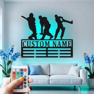 May include: A black metal wall decor featuring three cricket players in silhouette. The decor includes a customizable nameplate with the text "CUSTOM NAME".