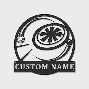 May include: Black metal wall art featuring a circular design with a central starburst shape and curved lines. The text "CUSTOM NAME" is displayed on a rectangular plaque below the design. The artwork is suitable for indoor or outdoor display.
