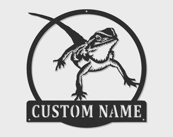 May include: A black metal wall sign featuring a circular frame with a detailed cutout of a bearded dragon lizard in the center. Below the circular frame is a rectangular banner with the text CUSTOM NAME in a bold, stencil-style font.
