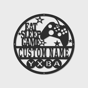 Può includere: Un'insegna da parete circolare in metallo nero con un design ritagliato di un controller per videogiochi, stelle e il testo EAT SLEEP GAME. Sotto, una barra orizzontale mostra il testo CUSTOM NAME, seguito da quattro cerchi contenenti le lettere Y, X, B e A.