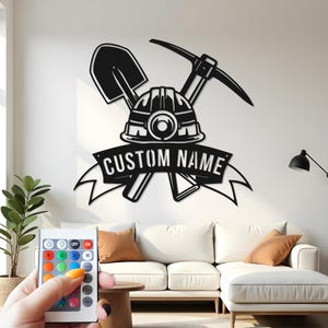 Könnte beinhalten: Schwarze Metall-Wandkunst mit einem Bergmannshelm, einer Spitzhacke und einer Schaufel. Das Design enthält ein Banner mit dem Text "CUSTOM NAME". Das Kunstwerk ist an einer weißen Wand über einem weißen Sofa angebracht.
