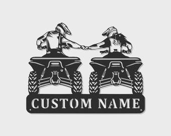 May include: A black metal wall art piece featuring two riders on ATVs facing forward, performing a fist bump. The design includes detailed tires, suspension, and helmets. Below the vehicles is a rectangular plate with the text CUSTOM NAME.