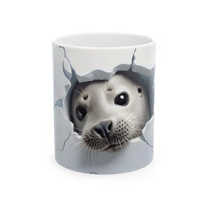 Può includere: Tazza in ceramica bianca con un'illustrazione realistica della testa di una foca che emerge da una superficie grigio chiaro strappata. La foca ha grandi occhi scuri e baffi. La tazza ha una finitura lucida.