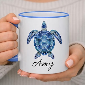 Puede incluir: Una taza de cerámica blanca con borde y asa azules, que presenta una ilustración detallada estilo acuarela de una tortuga marina azul centrada en el frente. Debajo de la tortuga, el nombre Amy está impreso en una elegante fuente cursiva negra.