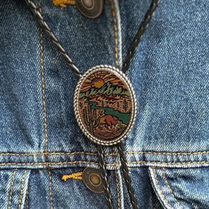 May include: Oval bolo tie with a desert landscape design. The silver-toned pendant features a colorful scene of mountains, a river, and a bison. The black leather cord is secured by a decorative clasp, set against a denim jacket.
