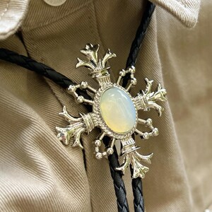 Op de afbeelding: Zilveren bolo stropdas met een ovale, opalescerende steen in het midden. De zilveren hanger heeft een sierlijk, kruisachtig ontwerp. Het zwarte gevlochten koord is te zien tegen een beige stoffen achtergrond.